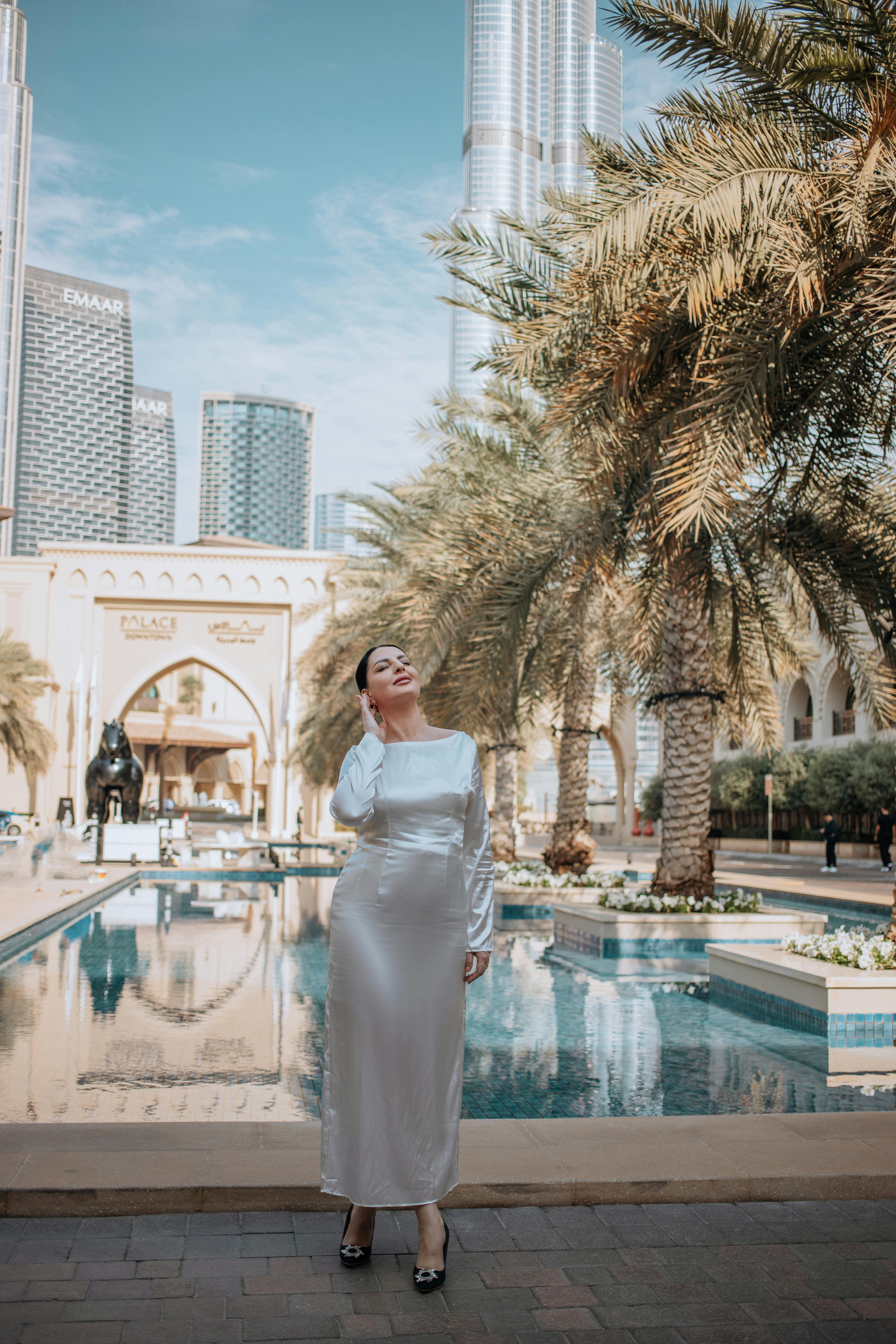 Zadarmo Žena v bielych šatách elegantne pózuje pri palmách v Dubaji a ukazuje modernú architektúru. Fotka z fotobanky