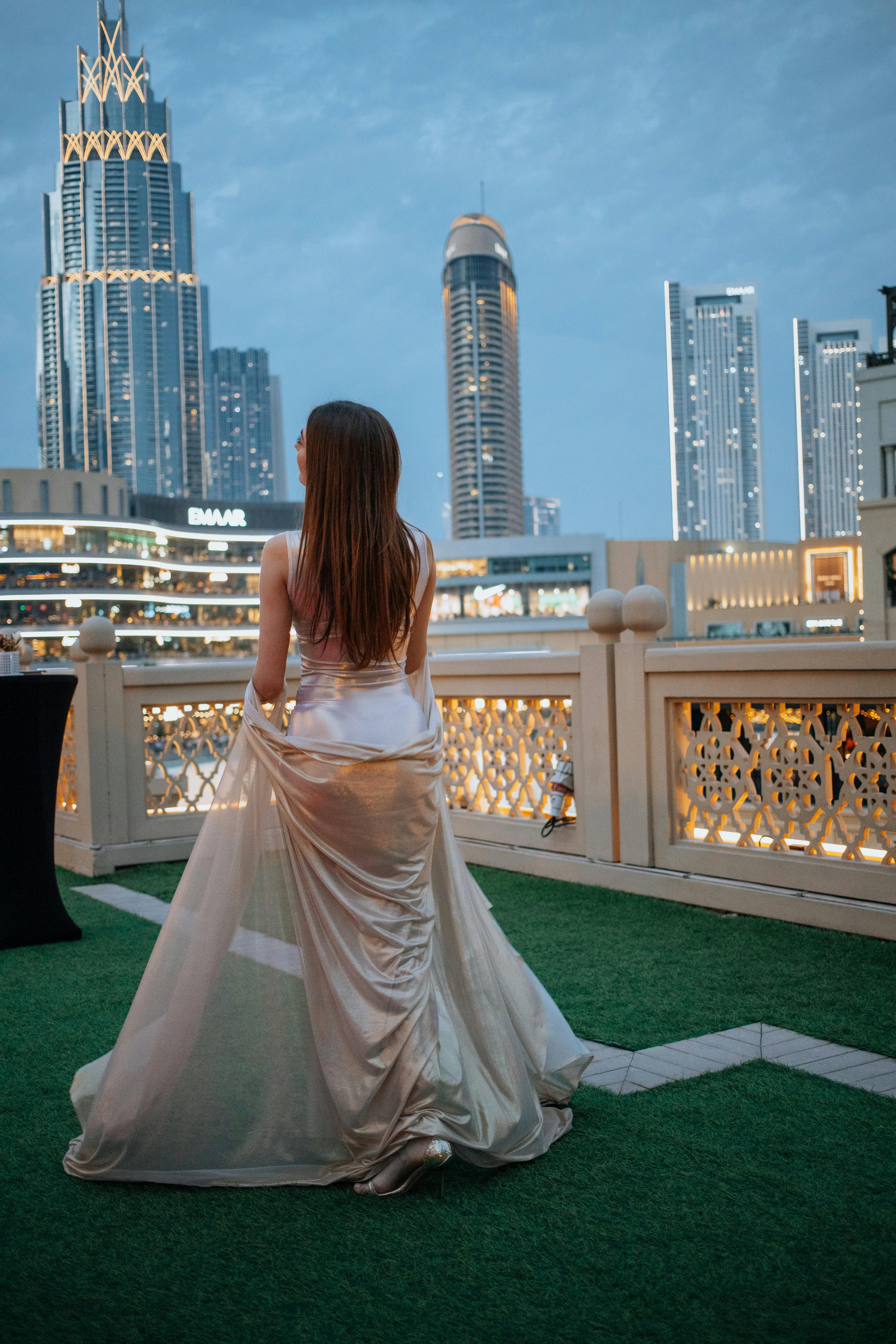 De franc Una dona amb un vestit elegant es troba en una teulada amb vistes a l'icònic horitzó de Dubai al capvespre. Foto d'estoc