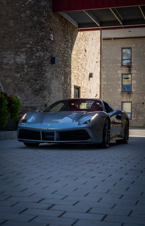 Free A sleek grey Ferrari sports car parked on a stone courtyard under a modern structure. Stock Photo