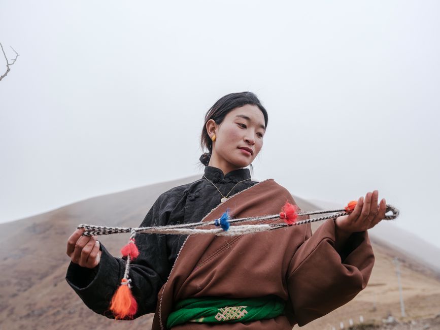 A young woman in traditional attire holds a handcrafted rope, showcasing cultural heritage in a rural mountain landscape.