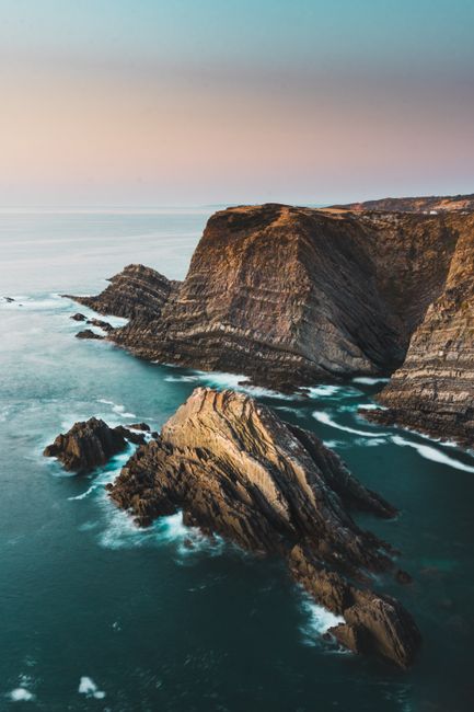 A breathtaking view of rugged cliffs and ocean waves in Beja District, Portugal during a serene sunset.