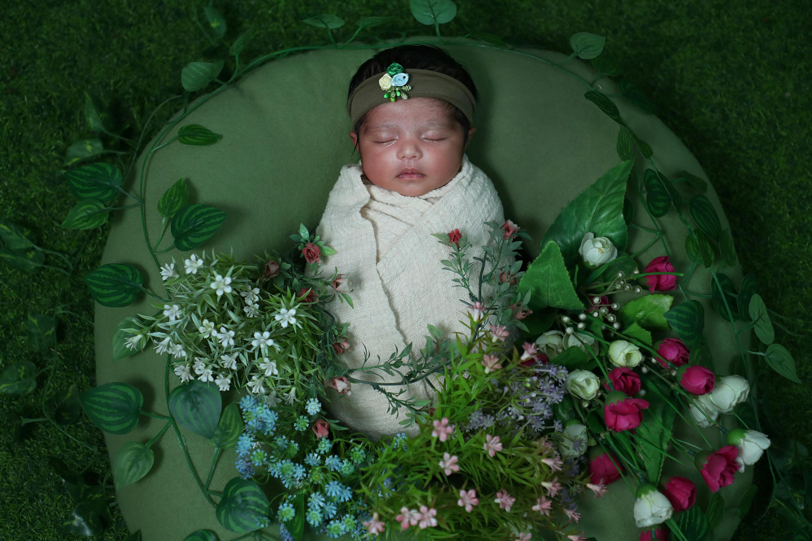 Adorable newborn baby wrapped in a soft blanket, surrounded by vibrant flowers, sleeping peacefully on a green bed.