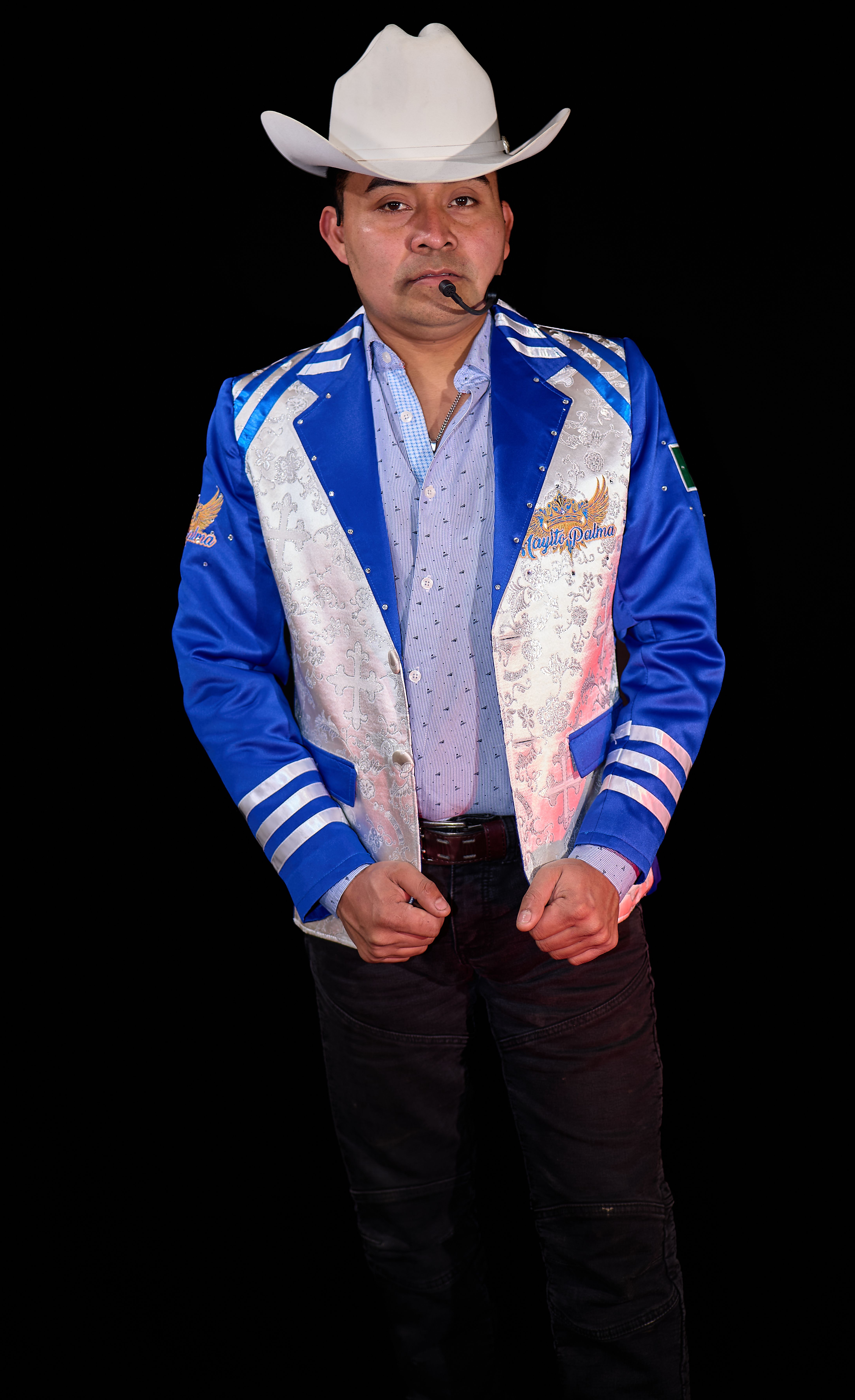 Man in blue blazer, cowboy hat, microphone on dark backdrop.
