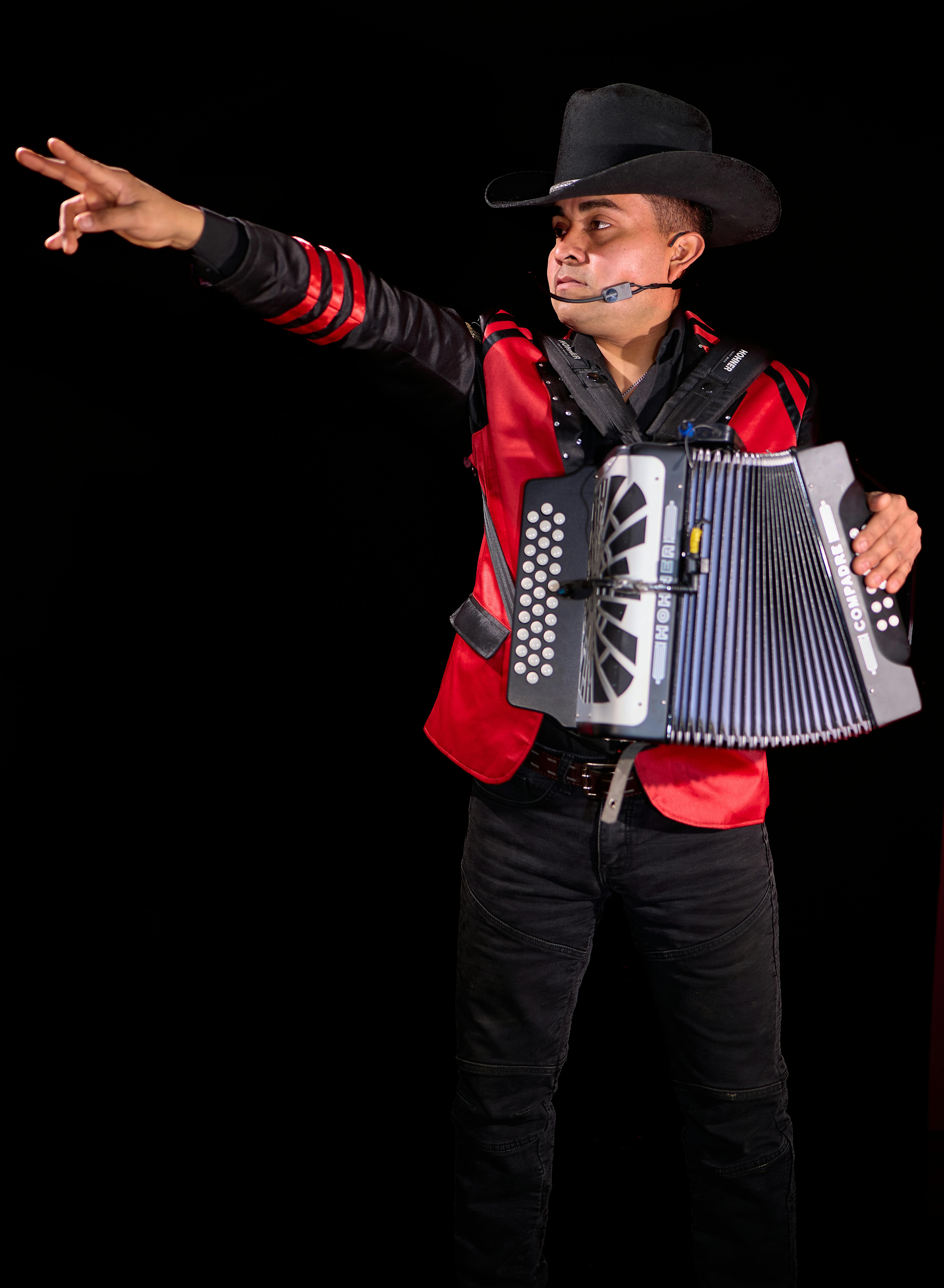 A lively musician entertains with an accordion, wearing a red jacket and cowboy hat, pointing dramatically.