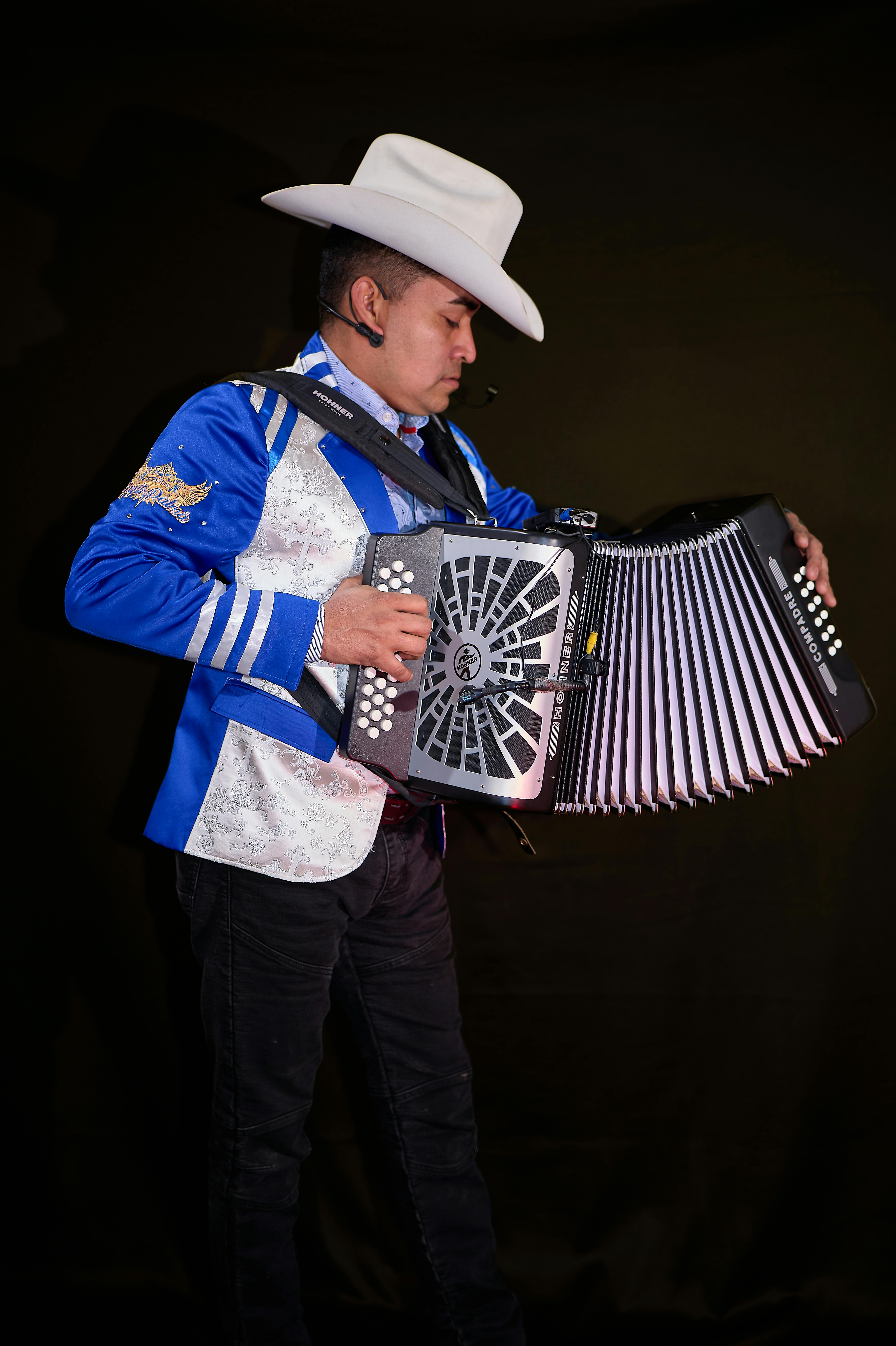 A musician in a blue costume and cowboy hat playing an accordion against a dark background.