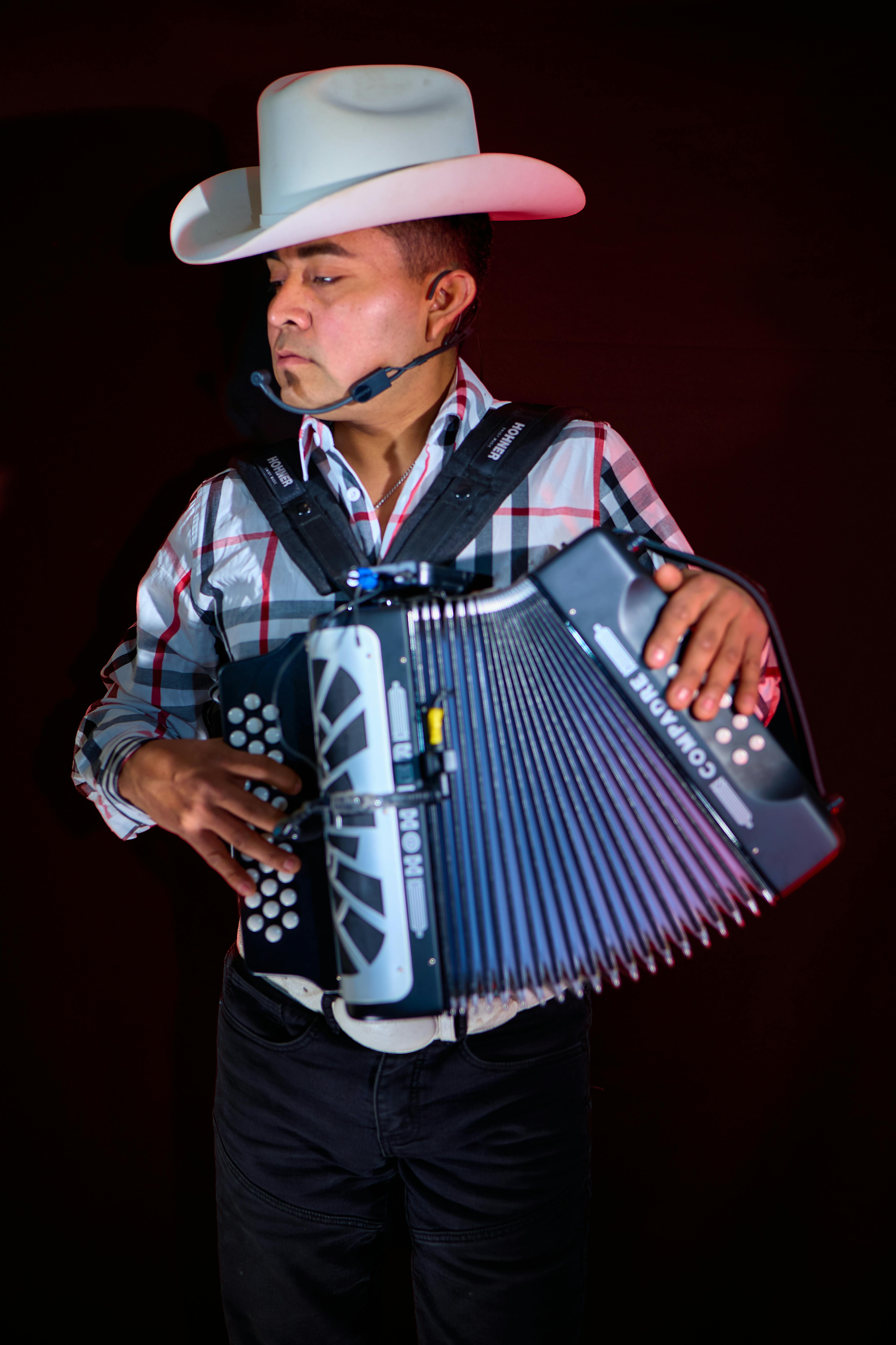 Adult male musician in cowboy hat plays accordion against dark backdrop.