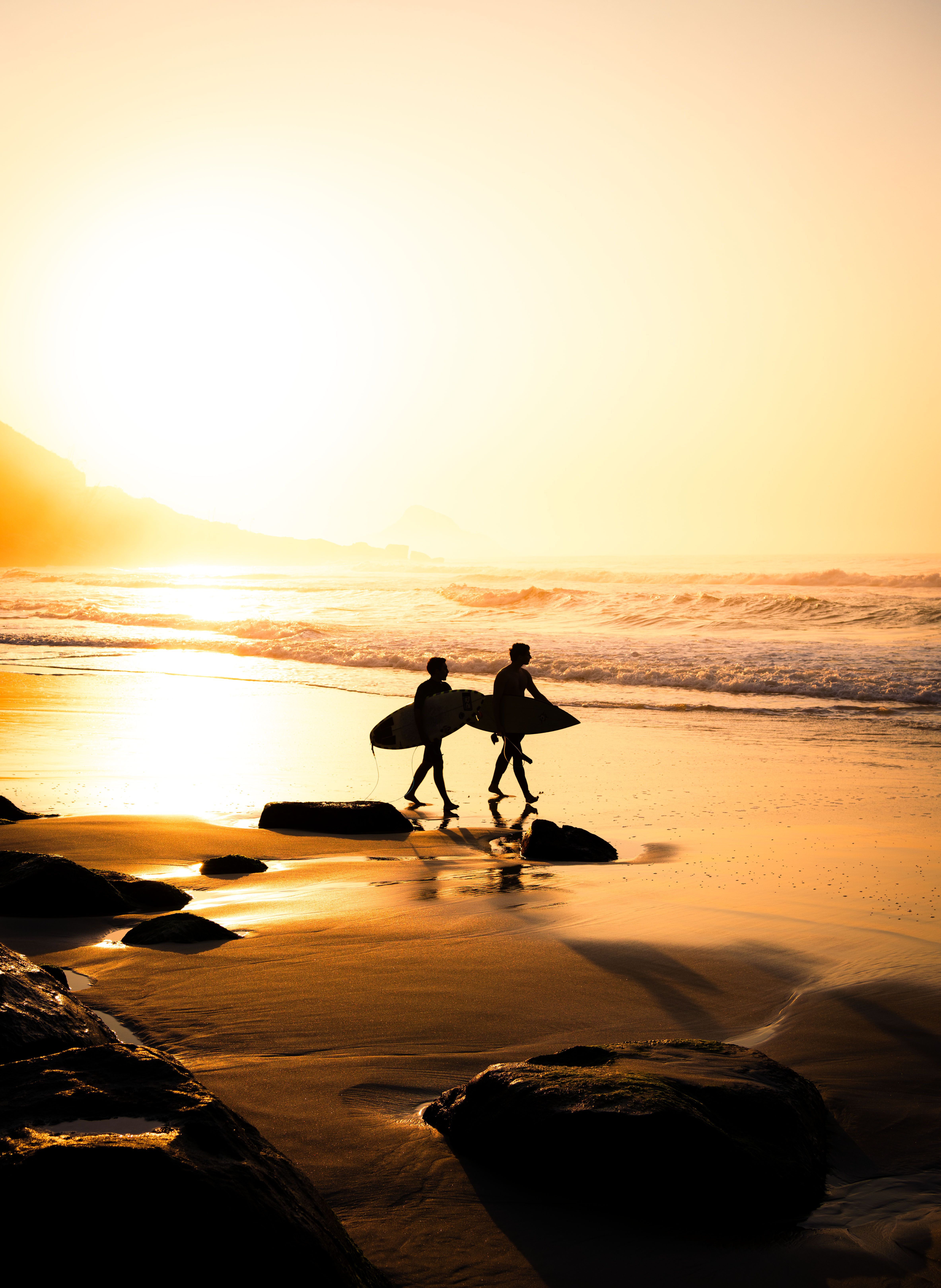 Two surfers walk along the shore with surfboards during a vibrant sunset, capturing the essence of beach life.