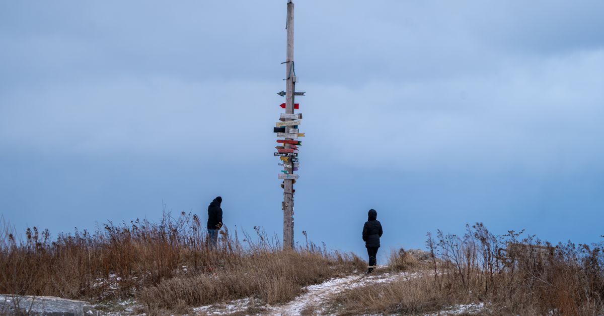 Two people in winter clothing explore a snowy landscape with a directional signpost under a cloudy sky.