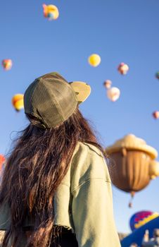 A woman in a cap gazes at colorful hot air balloons floating in the sky during a sunny day festival.