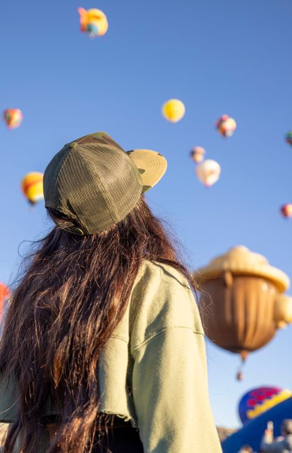 A woman in a cap gazes at colorful hot air balloons floating in the sky during a sunny day festival.