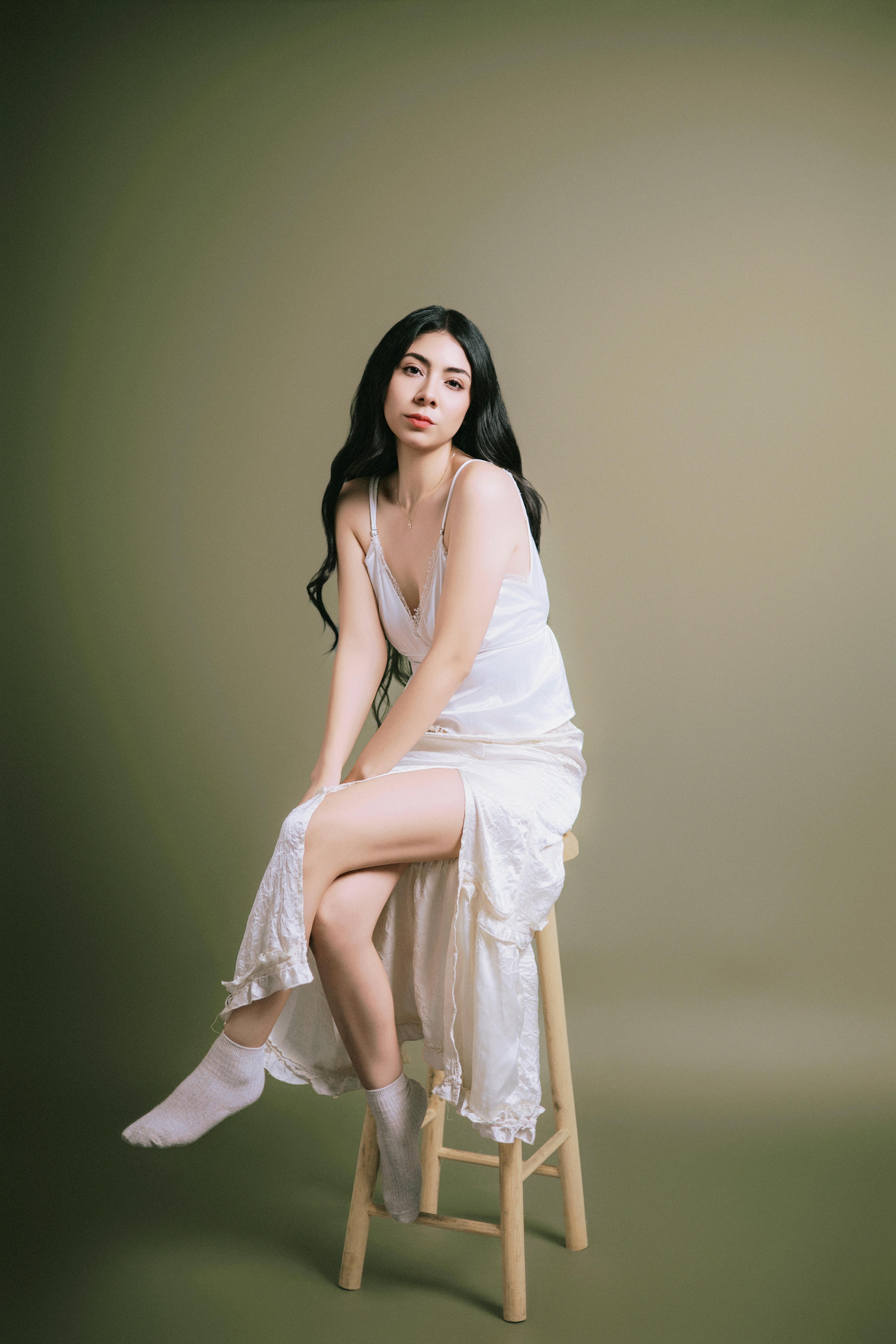 Graceful woman in a white dress sitting on a stool against a muted backdrop.