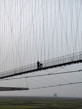 A cyclist pushes their bike across a high suspension bridge on a foggy day.