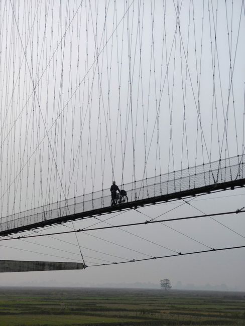 A cyclist pushes their bike across a high suspension bridge on a foggy day.