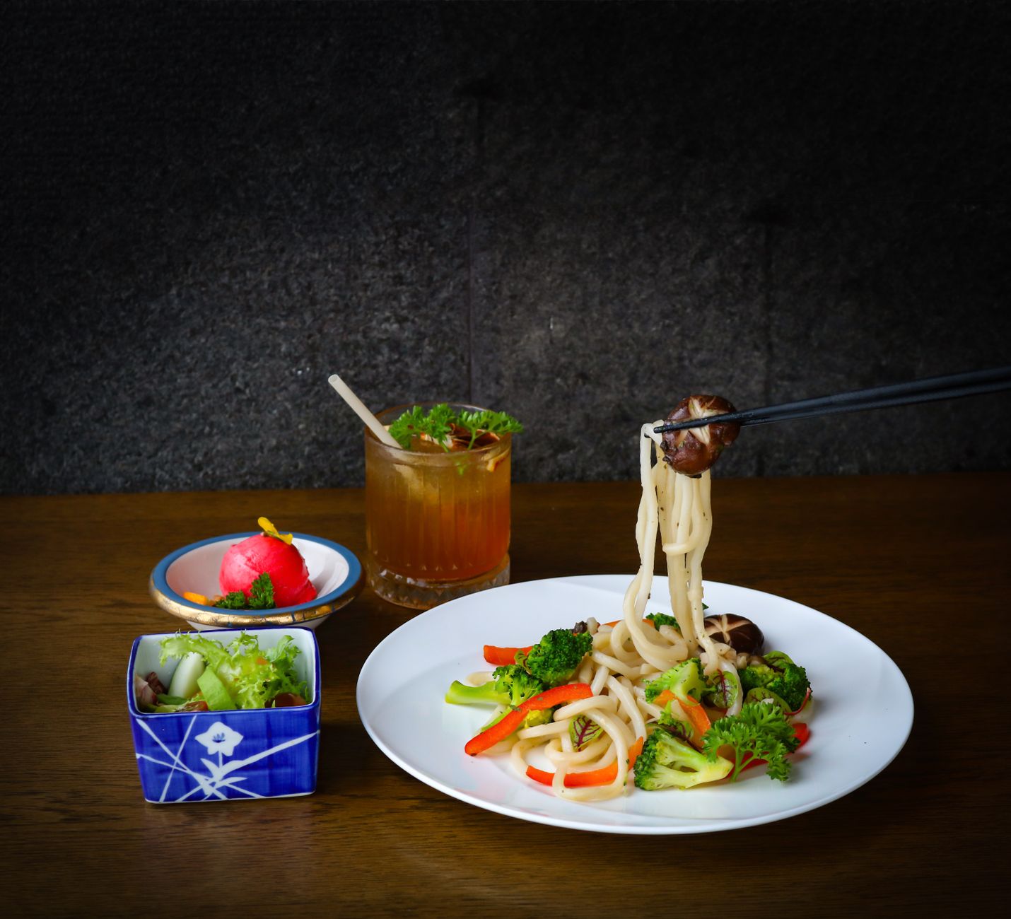 Delicious udon noodle dish with vegetables, accompanied by salad and a refreshing drink, presented in an artistic setup.