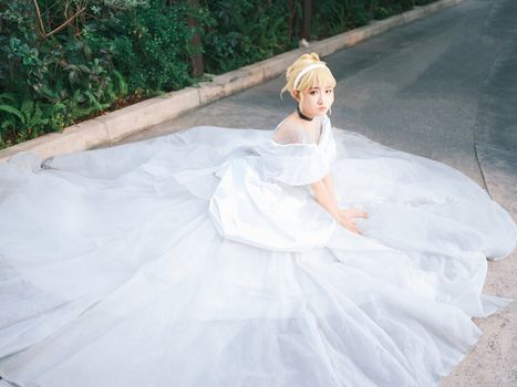 A woman in an elegant white gown sitting on an outdoor pathway, surrounded by greenery.