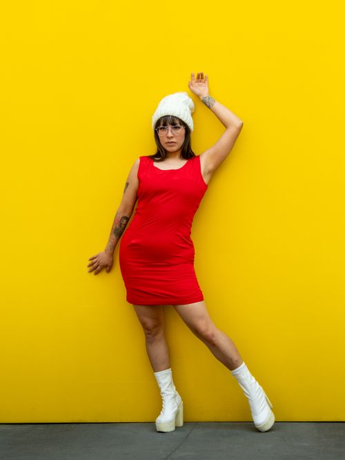 Free Adult woman in red dress and white beanie poses stylishly against a vibrant yellow wall. Stock Photo