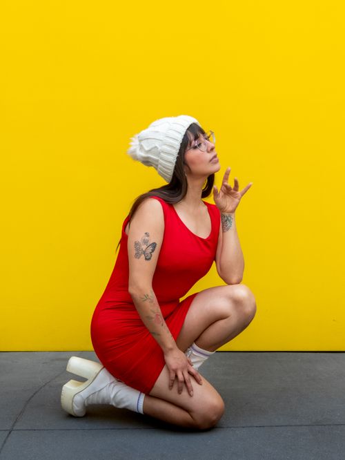 Free A fashionable woman kneels in a red dress and white beanie against a vibrant yellow backdrop. Stock Photo