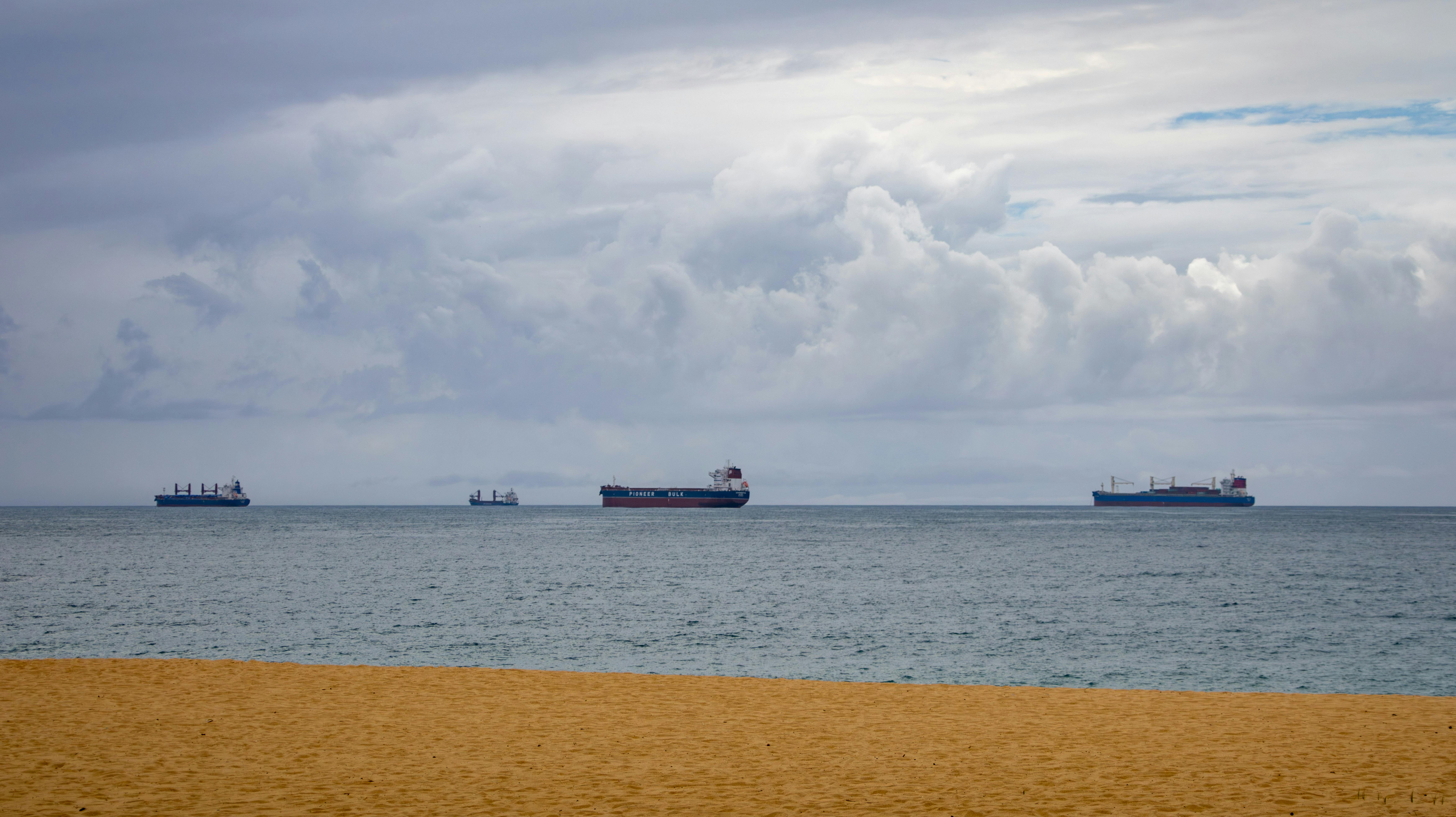 Four cargo ships float on the ocean, viewed from a sandy beach under a cloudy sky.