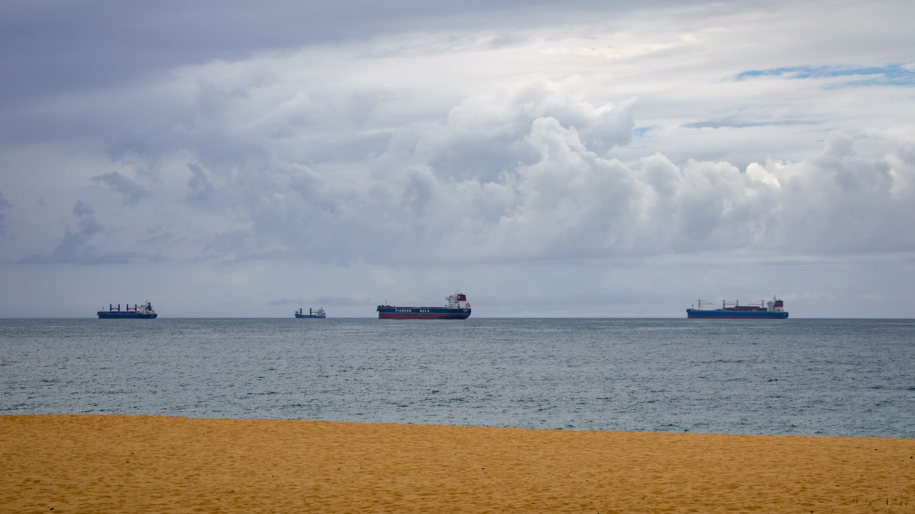 Four cargo ships float on the ocean, viewed from a sandy beach under a cloudy sky.