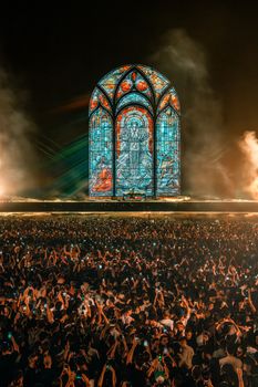 Massive crowd enjoying a nighttime concert with a stunning stained glass display on stage.