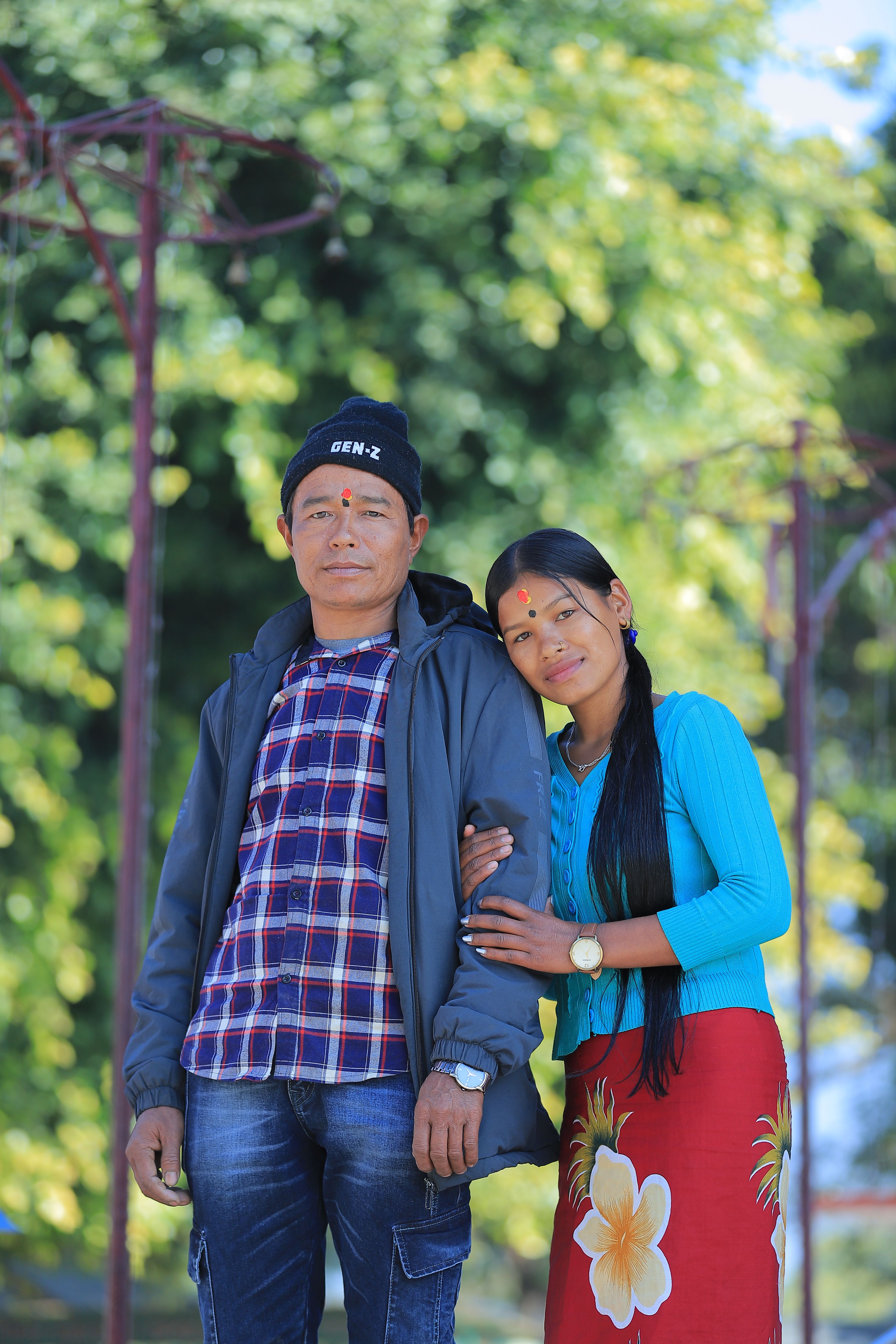 A smiling couple poses together outside on a bright sunny day, surrounded by nature.