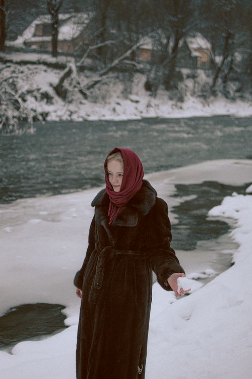 Free Woman in a coat and scarf standing by a snowy riverbank on a winter day. Stock Photo