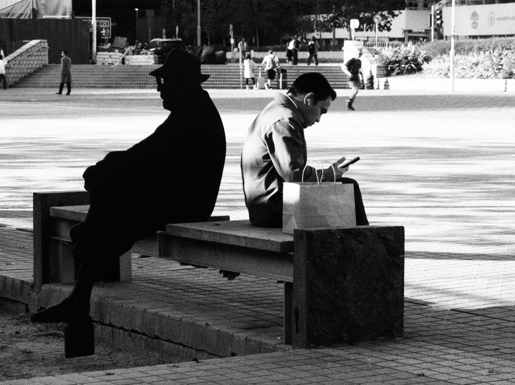 Black and white photo of two people on a bench in Hong Kong, one using a smartphone.