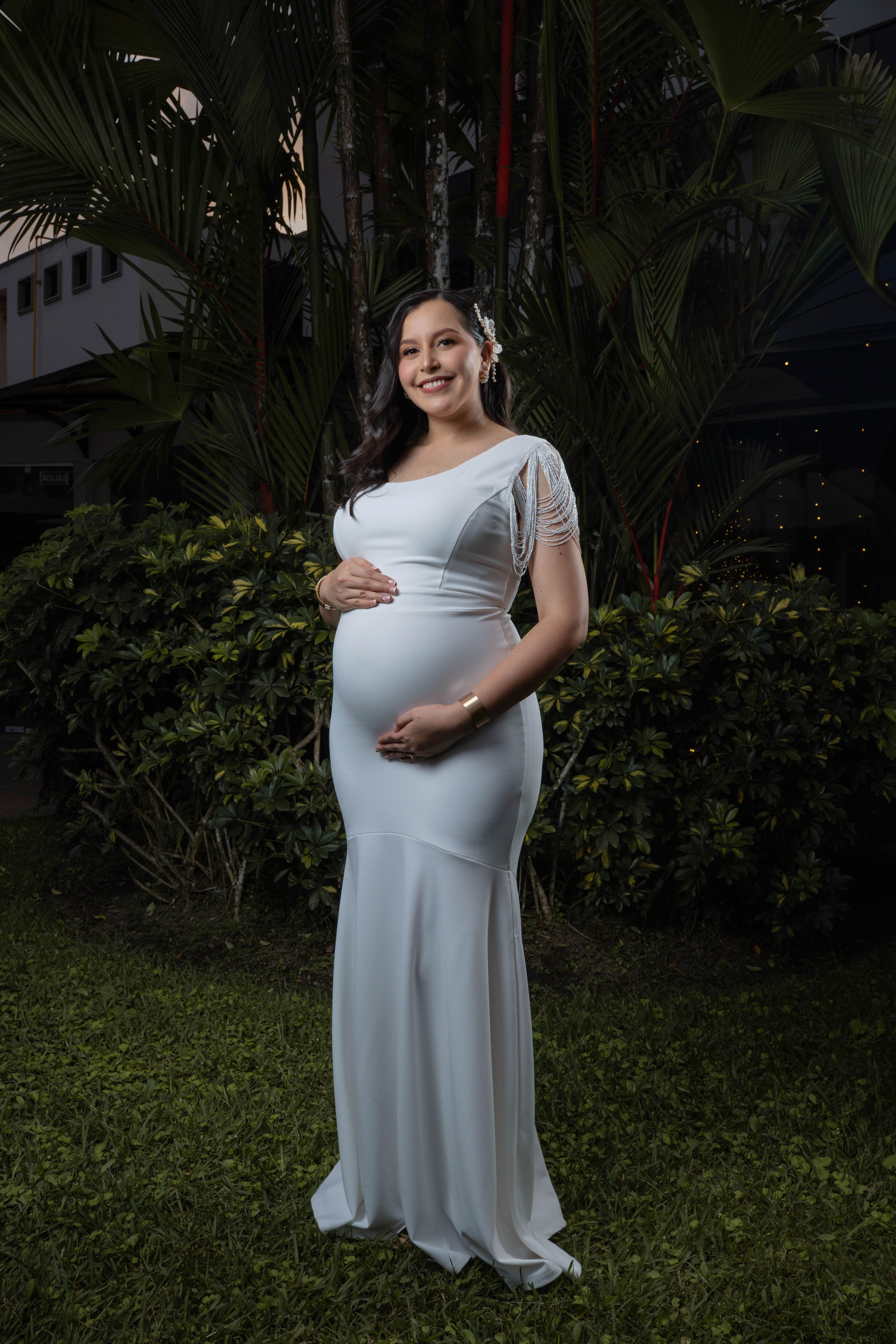 A pregnant woman in a white gown poses in a lush outdoor garden at twilight.