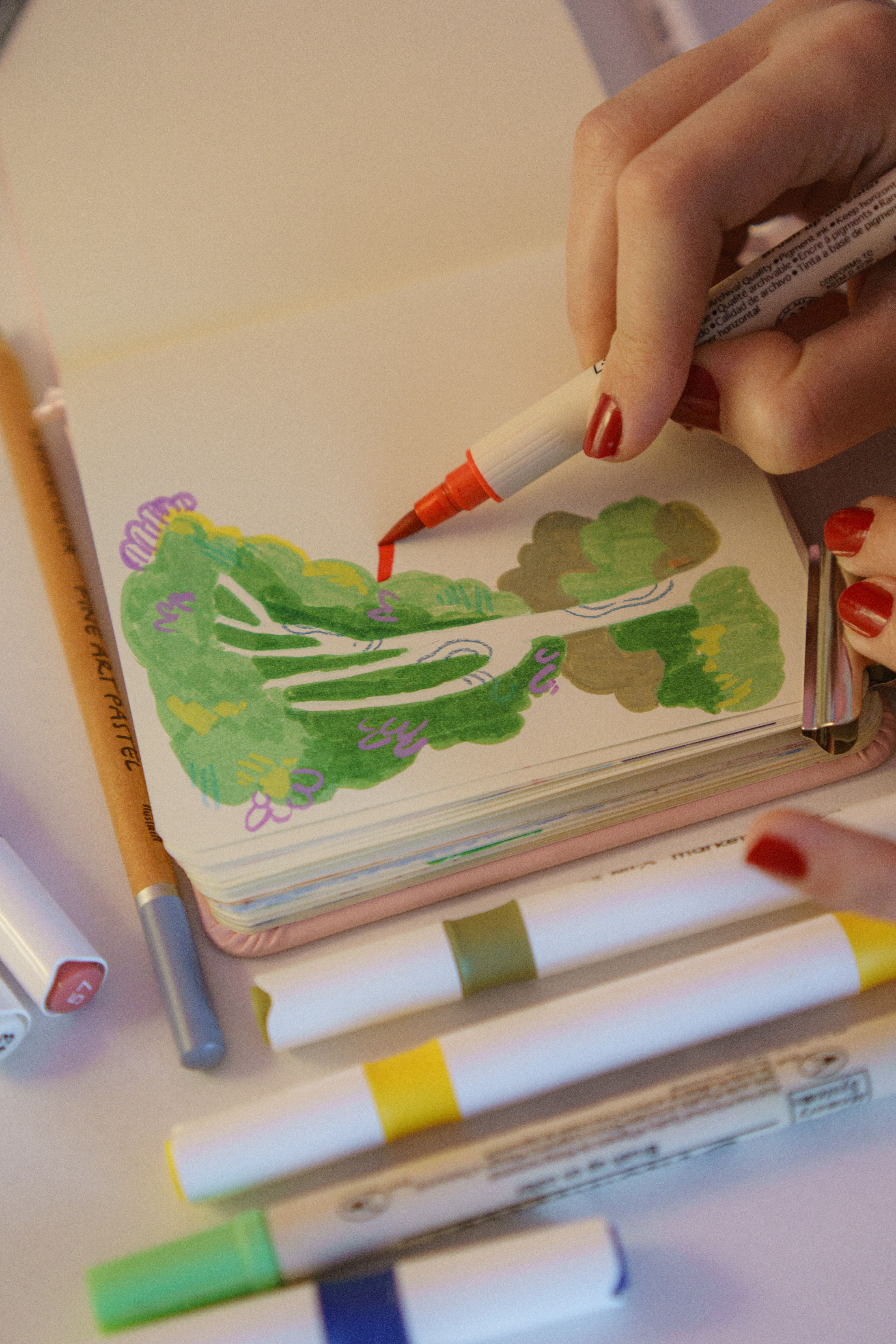 A person drawing vibrant greenery with markers in a sketchbook, featuring creative expression.