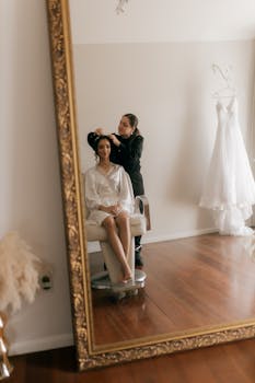 A bride getting her hair styled before the wedding, reflected in a large ornate mirror.