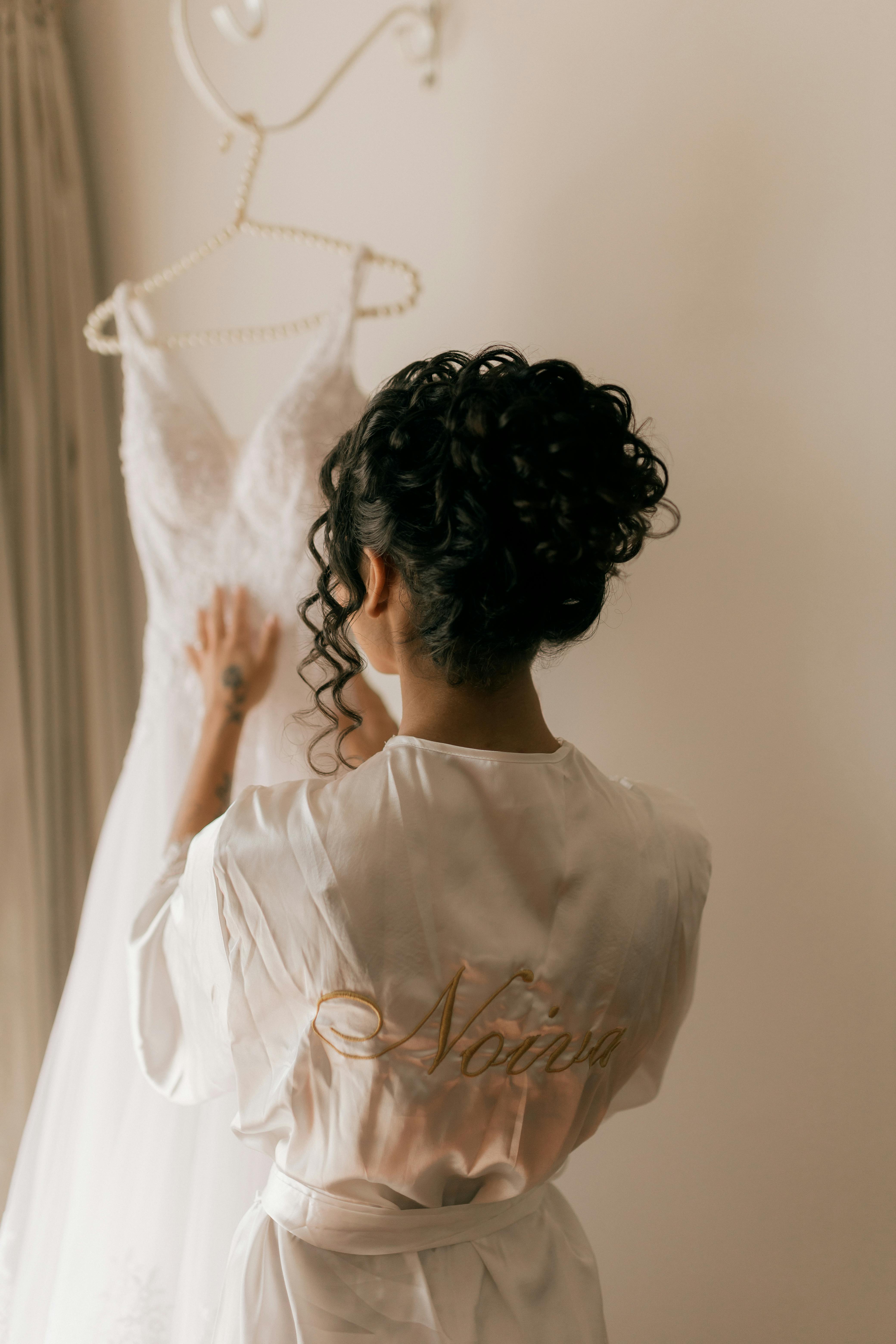 Bride in silk robe admires her elegant wedding dress hanging in bright room.