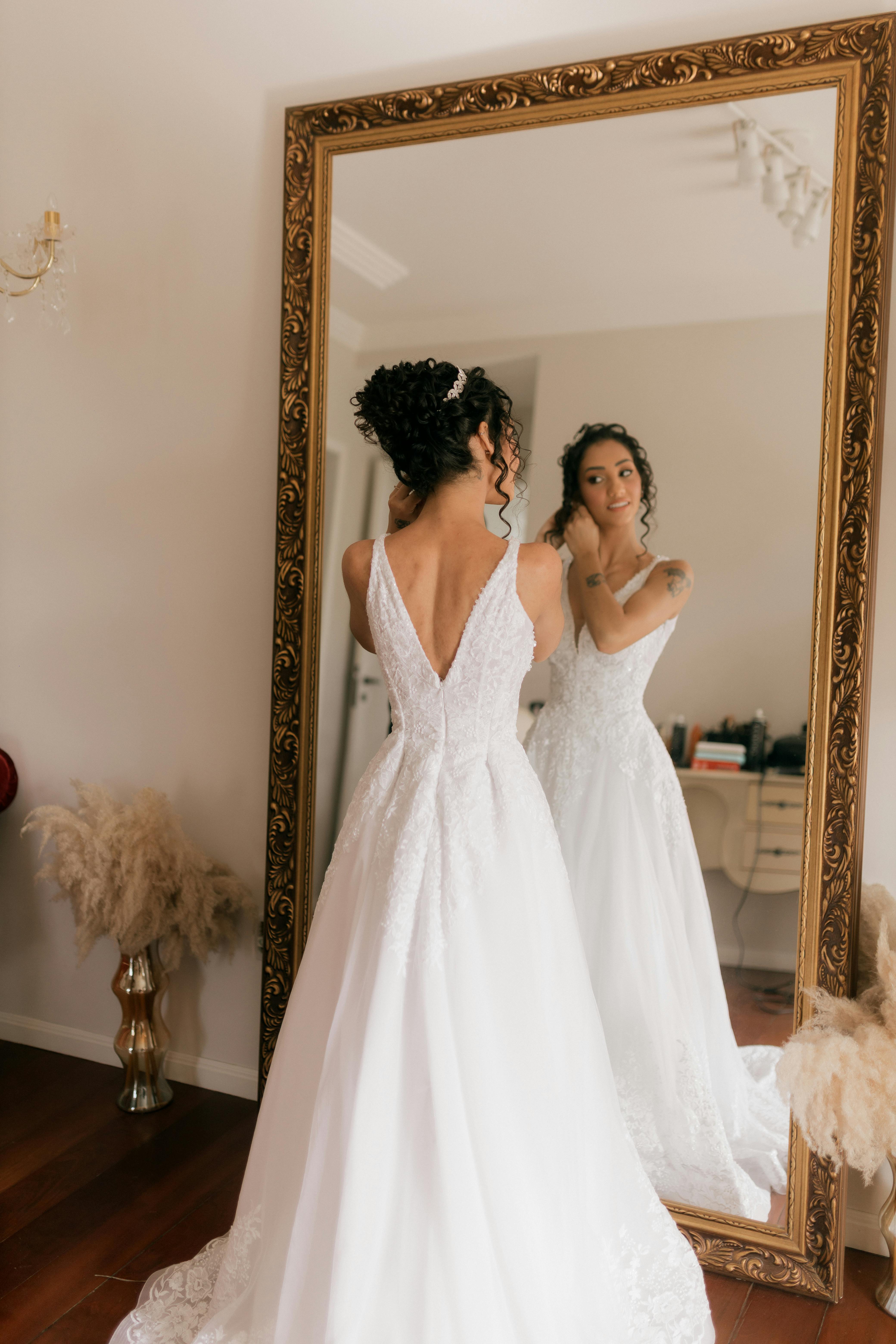 Bride in wedding dress looking at mirror reflection indoors.