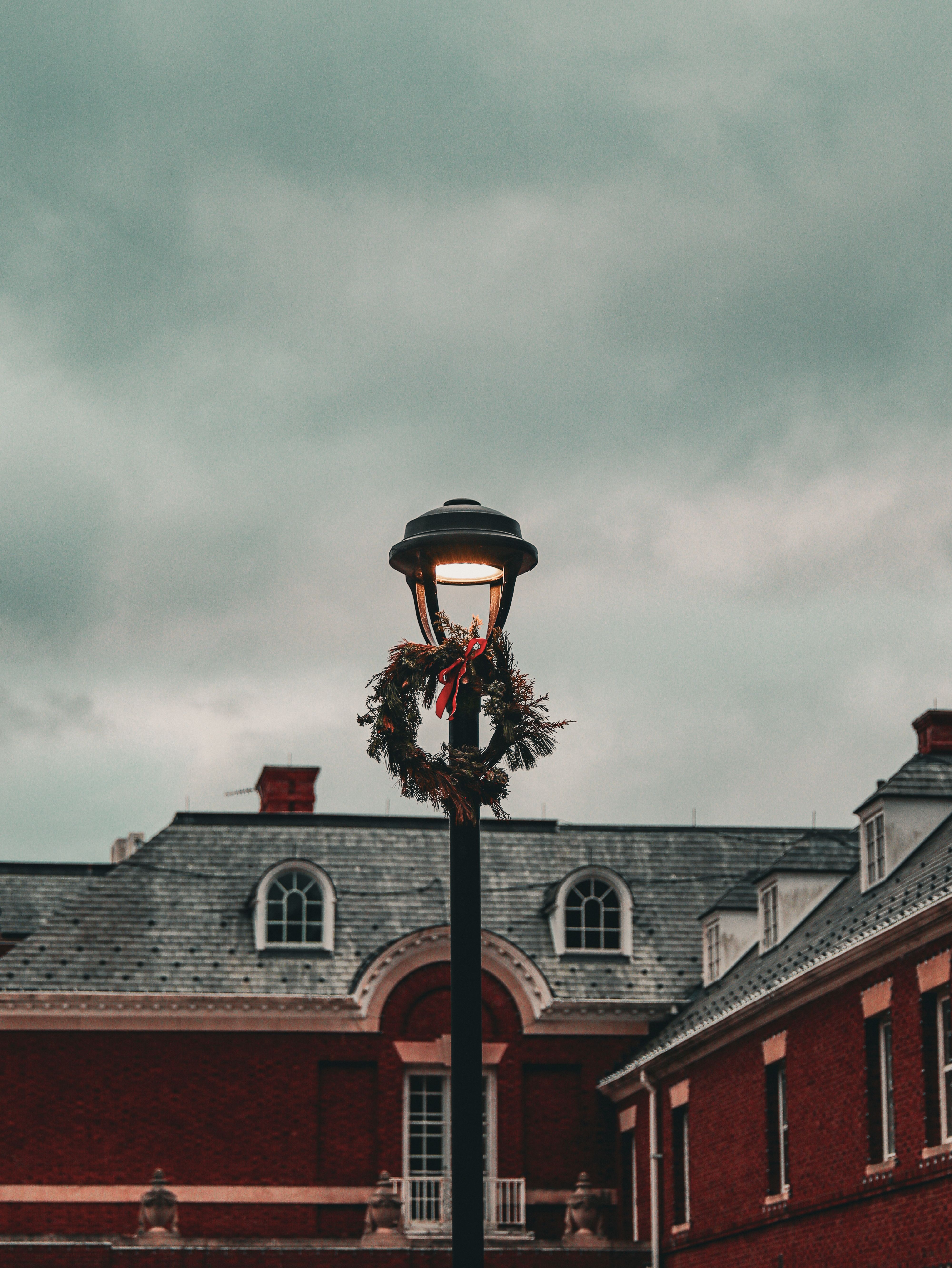 Vintage lamppost adorned with a Christmas wreath against an old building under cloudy skies.