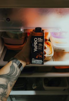 Tattooed arm holding a chocolate protein drink in an open refrigerator.