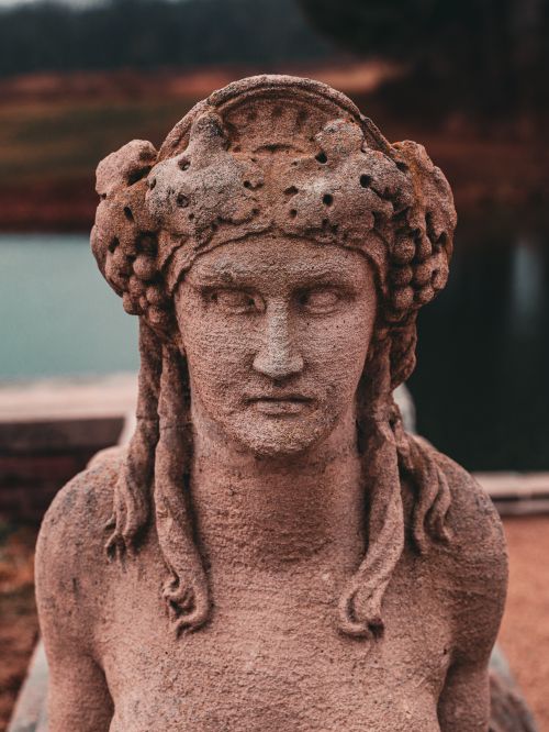 Free Close-up of a weathered ancient stone sculpture of a woman with intricate headpiece near a serene water body. Stock Photo