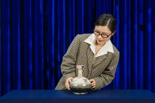 Woman examining a decorative vase in an elegant setting with a blue curtain backdrop.