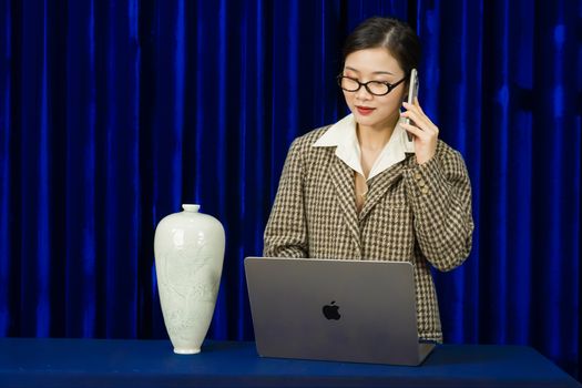 Businesswoman in a plaid jacket using phone and laptop beside a decorative vase against blue curtain.