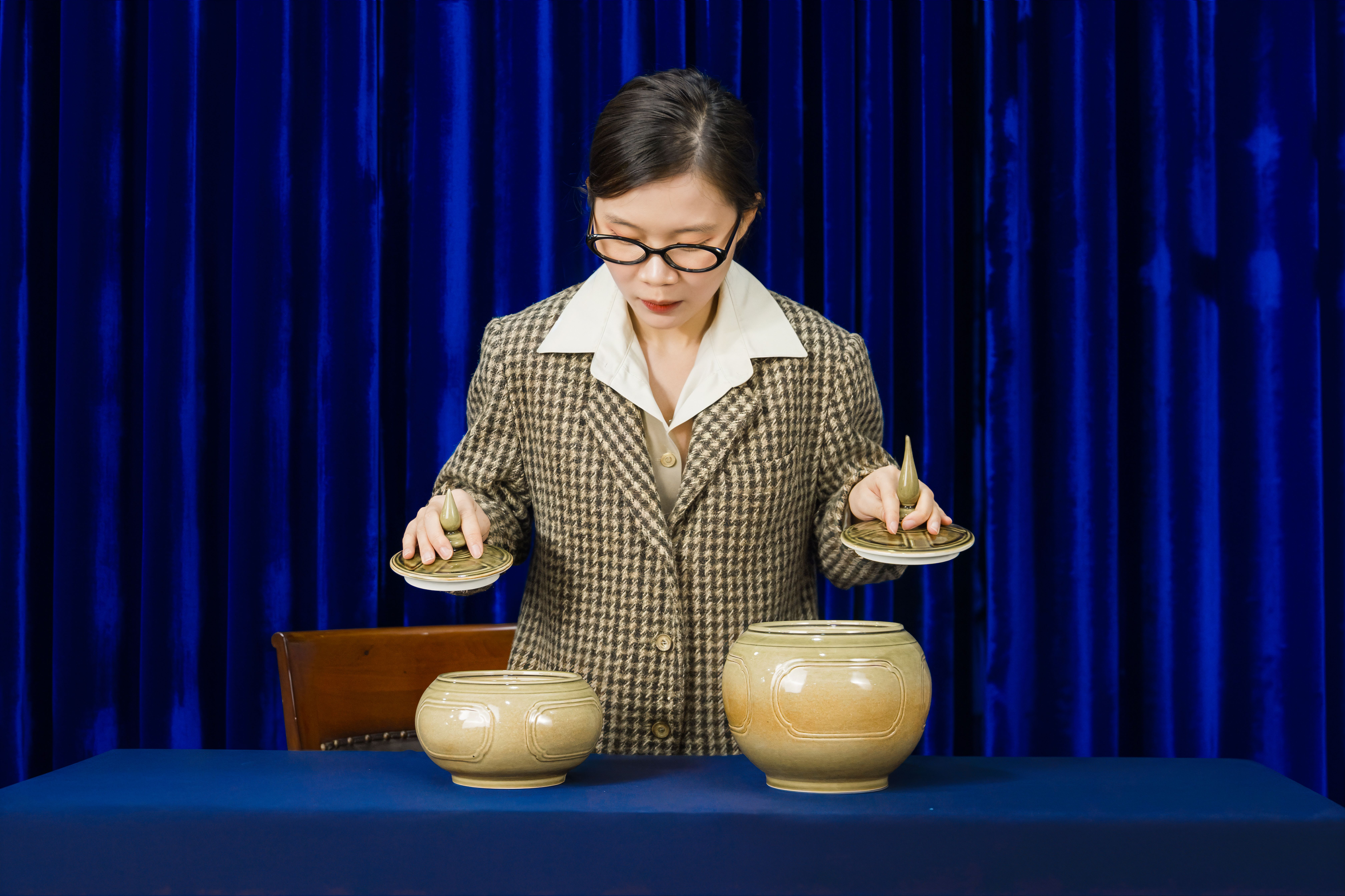 Woman in suit arranging ceramic vases on a blue table indoors with a vibrant curtain backdrop.