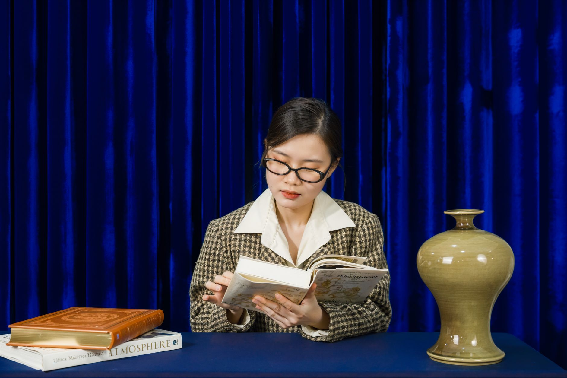 A young woman reading a book at a table with a classic decor vase; set against a dark blue curtain.