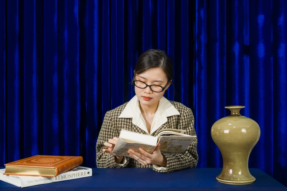 A young woman reading a book at a table with a classic decor vase; set against a dark blue curtain.