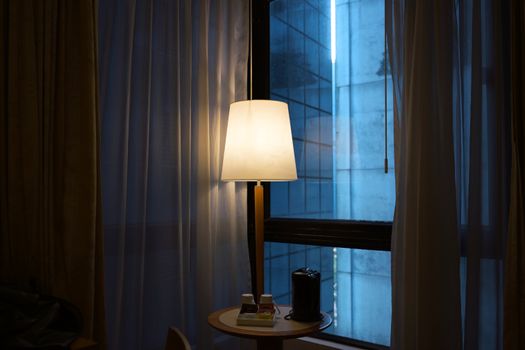 Warmly lit room with a glowing lamp and urban cityscape view through a window.