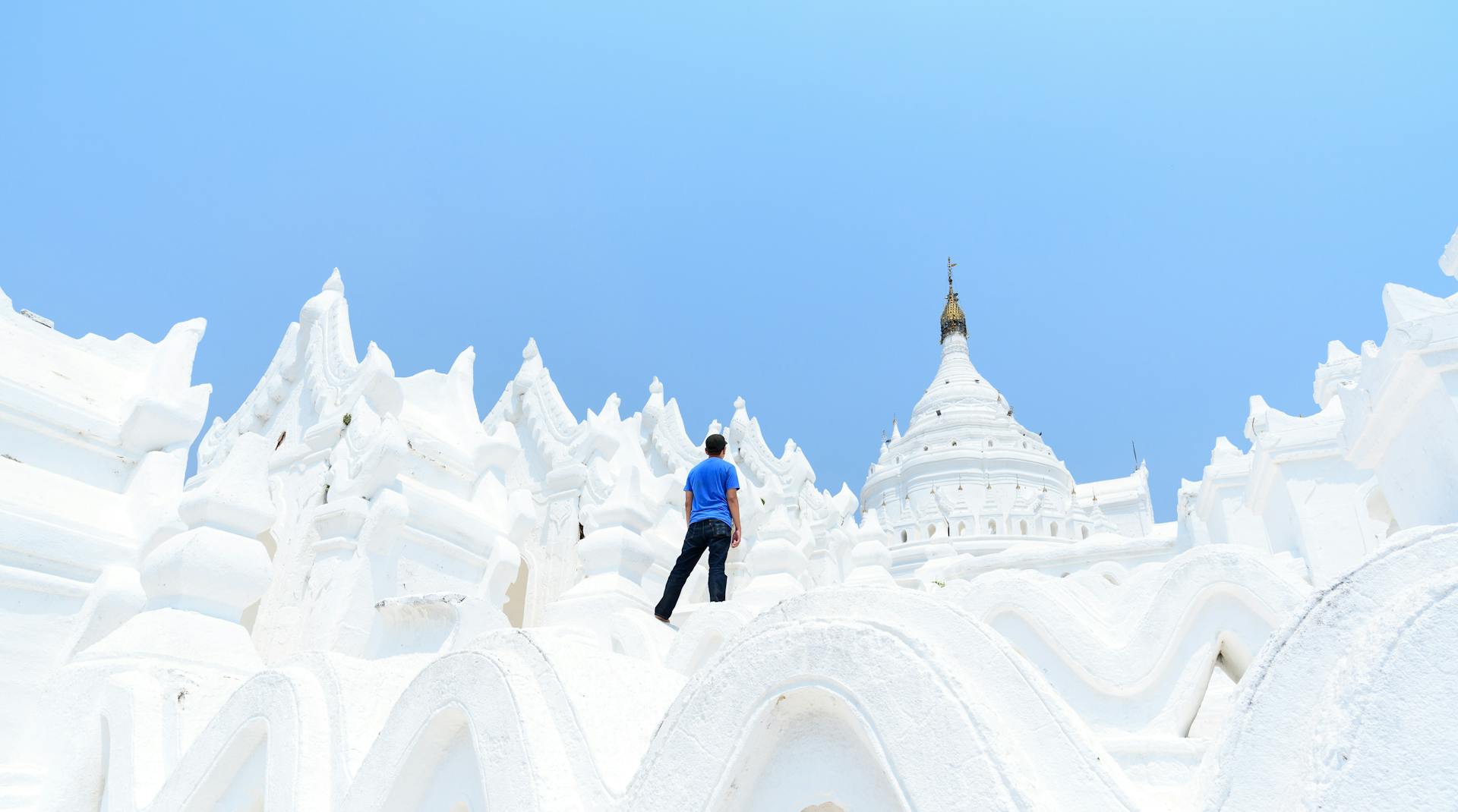 A man in blue ascends the white Hsinbyume Pagoda under clear skies in Myanmar.