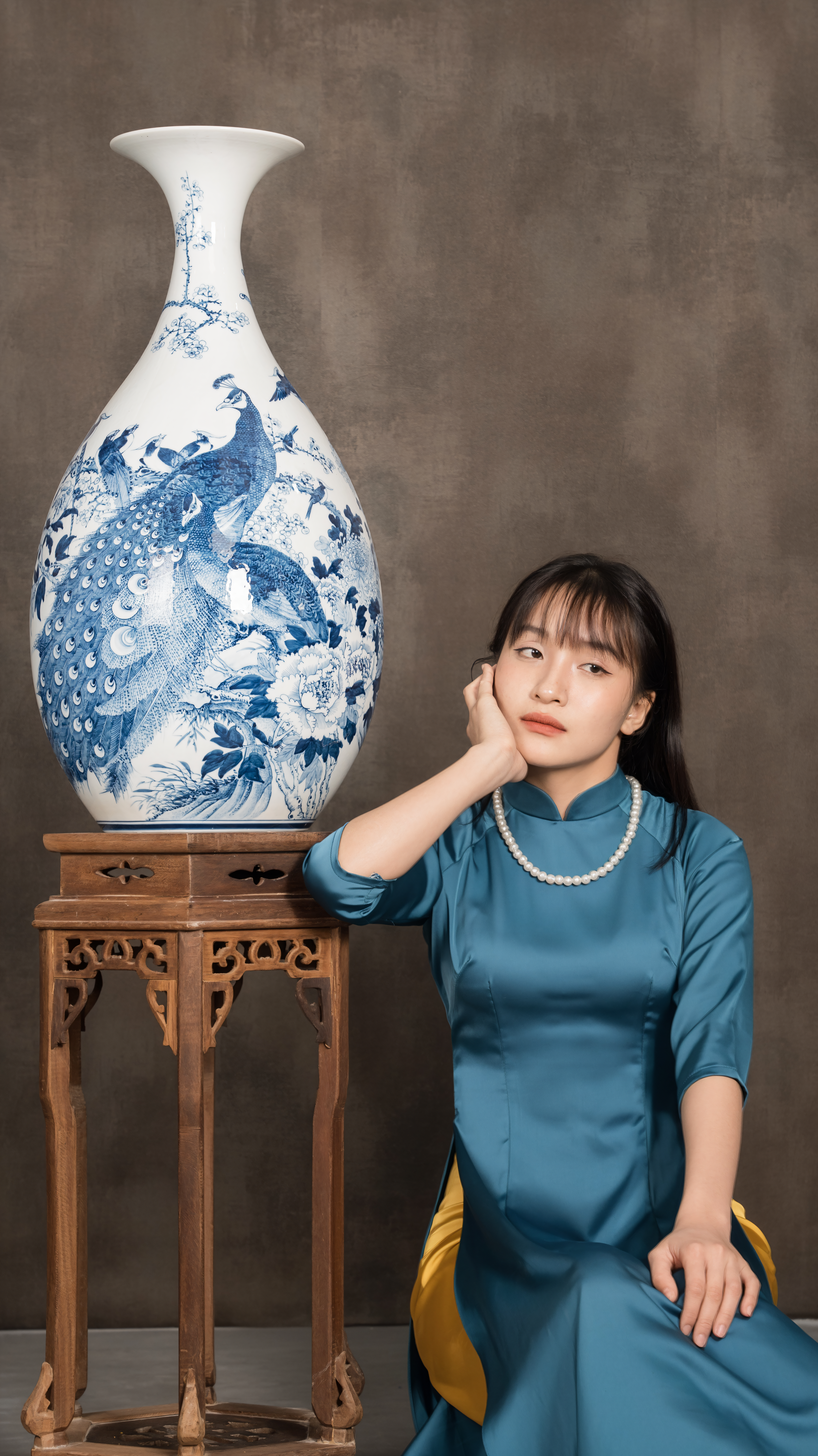 A Vietnamese woman in a teal dress poses beside a traditional ornate vase, blending modern elegance with cultural artistry.