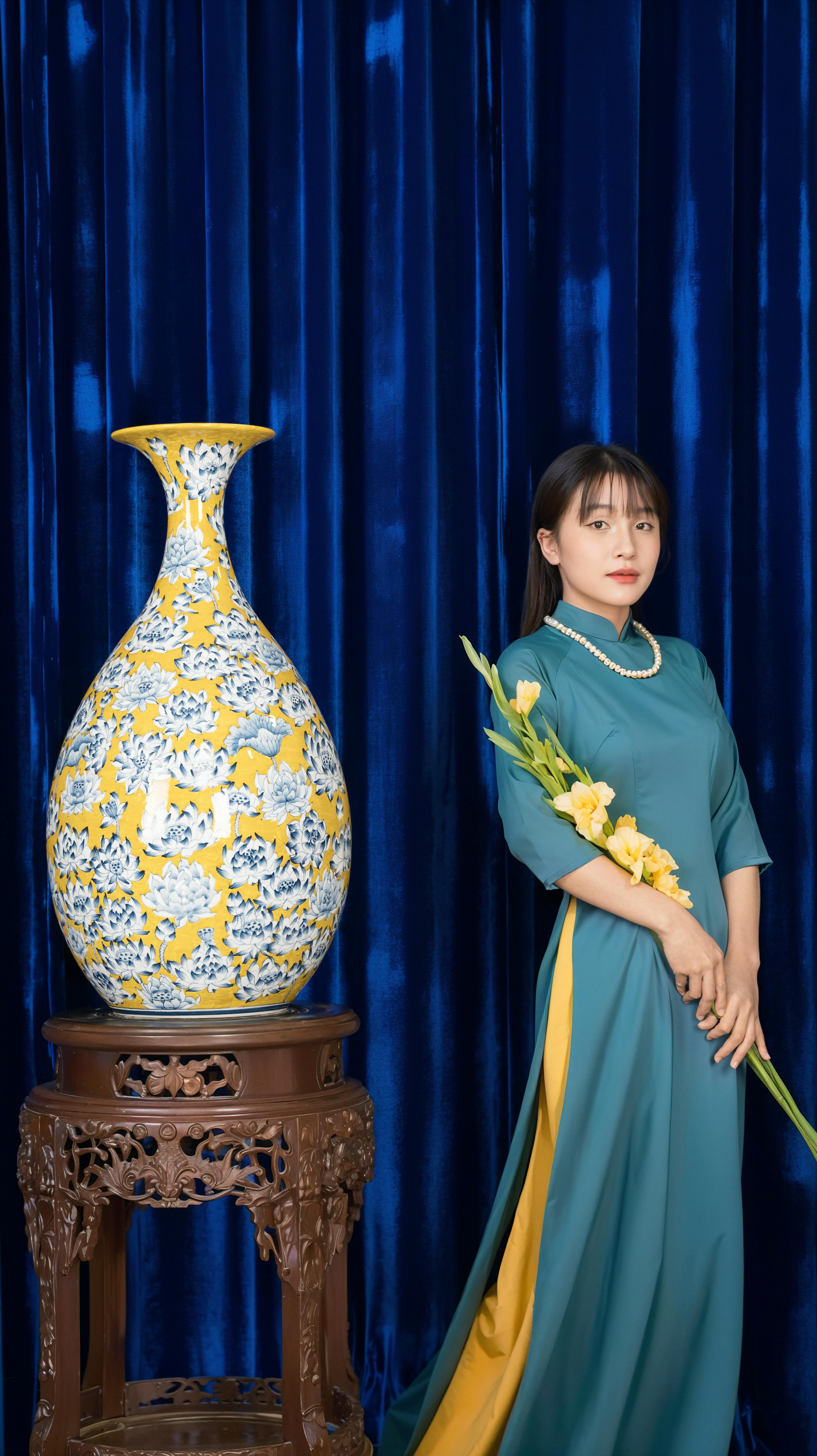 A woman in traditional dress poses with a Vietnamese vase and flowers against a blue backdrop.