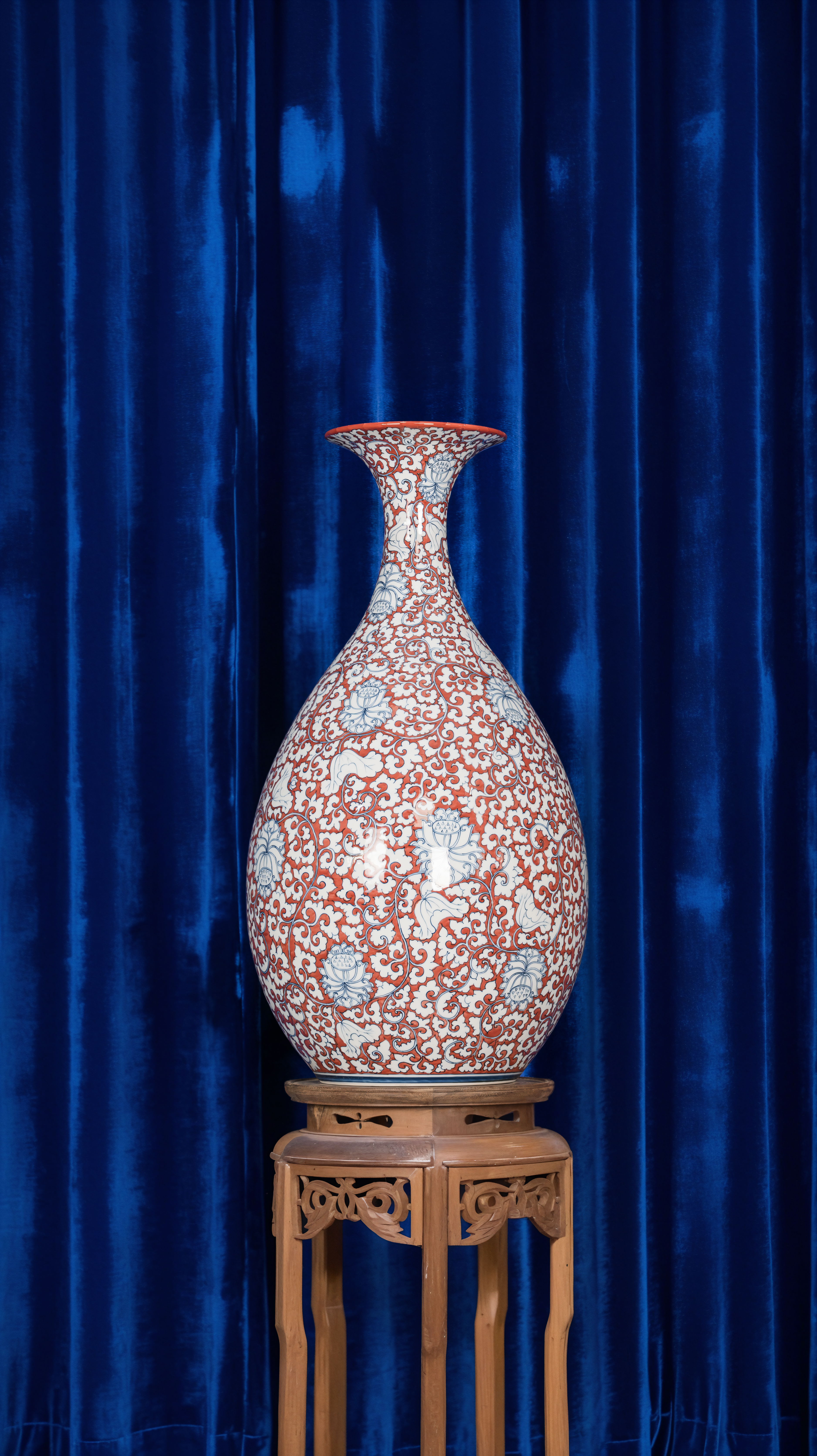 A beautifully ornate vase displayed on a wooden stand against blue velvet curtains.