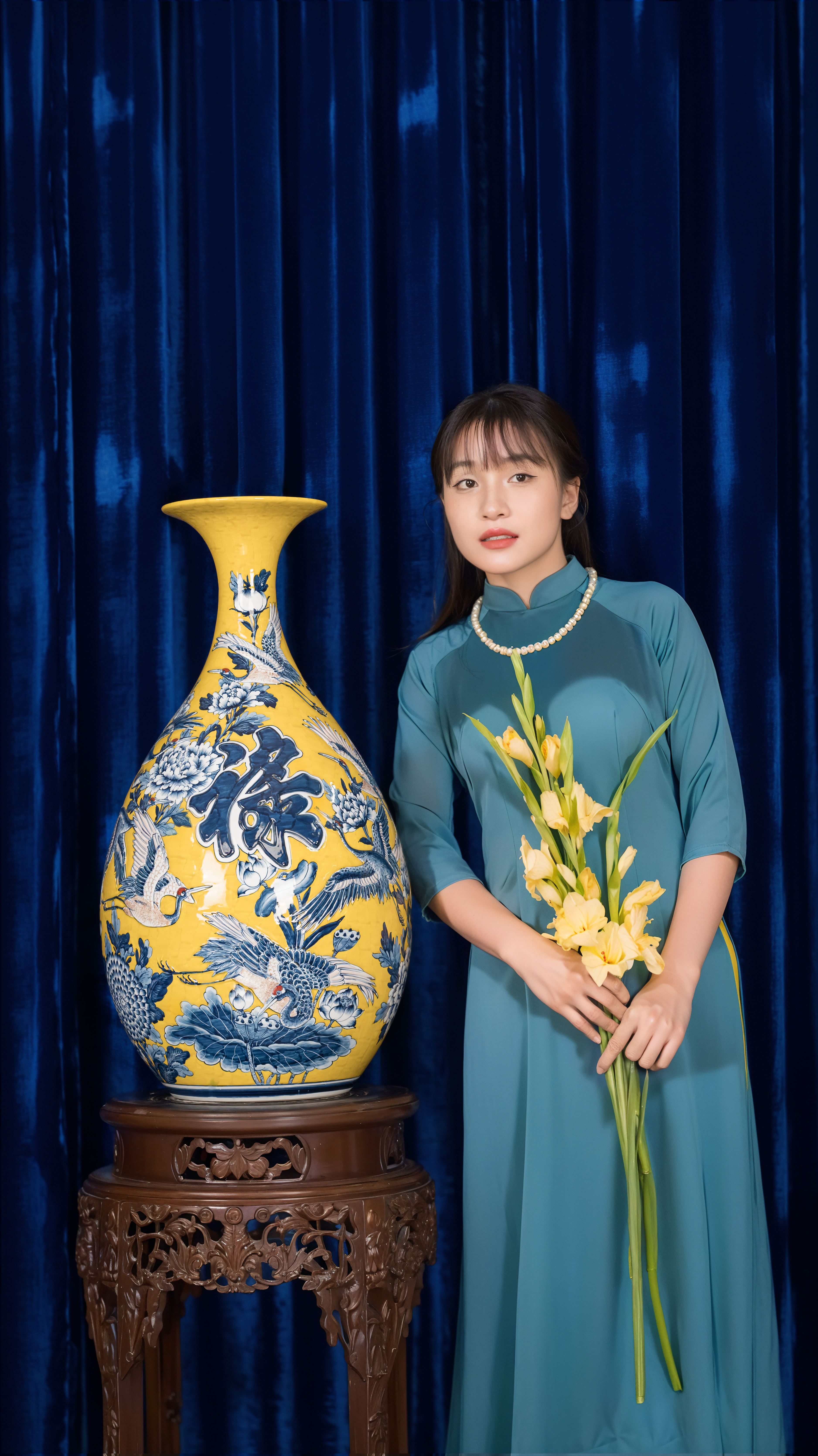 Woman in traditional attire next to ornate vase, holding yellow flowers.
