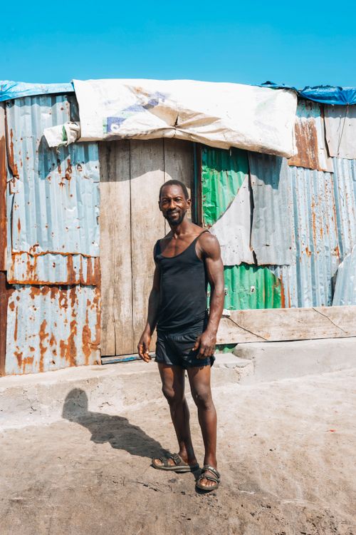 Free An adult man in casual attire stands outside a corrugated iron house under clear sky in Monrovia, Liberia. Stock Photo