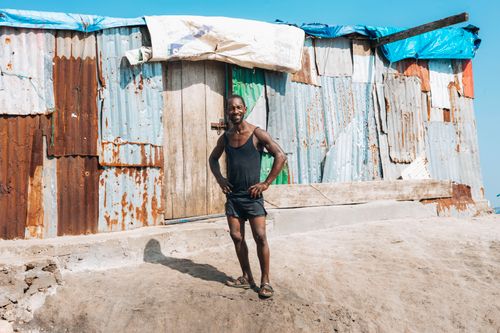 Free Smiling man stands confidently by a colorful metal shack under bright sunlight. Stock Photo