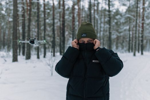 A person in winter clothing stands in a snowy forest with a flying drone nearby.