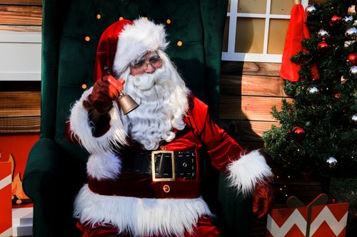 Free Santa Claus sitting on a green chair with a bell, next to a decorated Christmas tree. Stock Photo